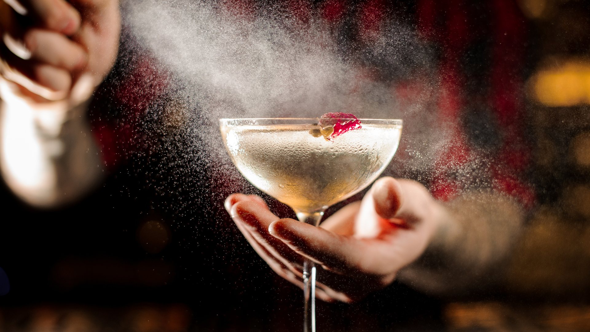 A bartender spraying a fine mist from an atomizer over a coupe held in one hand