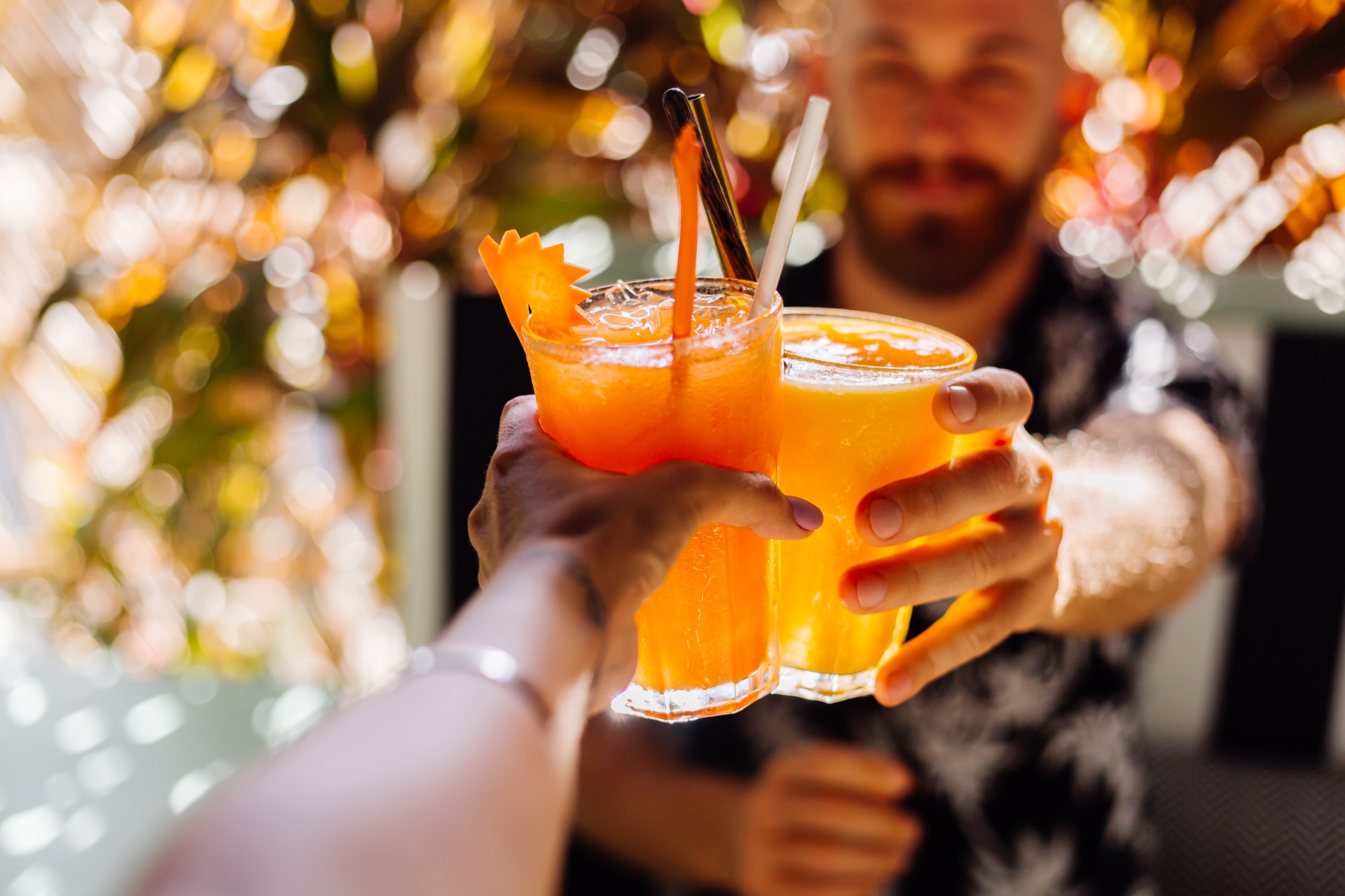 Two people clinking tall orange cocktails together in a sunlit cafe