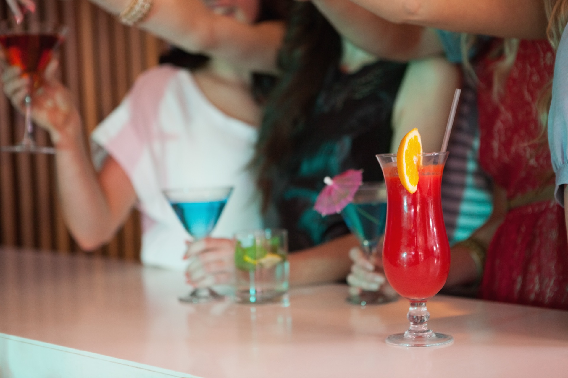 A lineup of colorful cocktail entries on a bar with friends behind