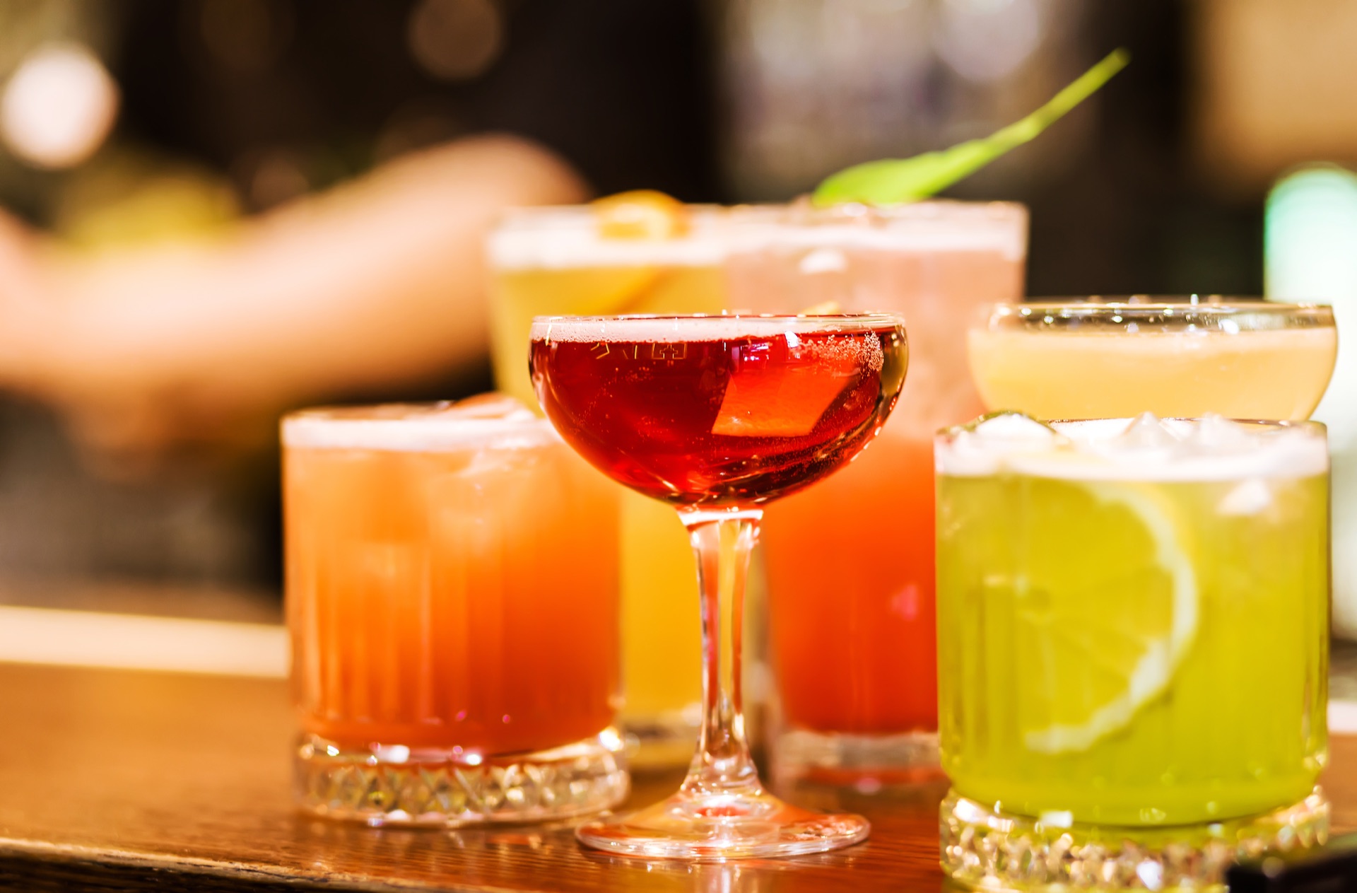 Six cocktails lined up on a bar counter, each a different colour and glass shape