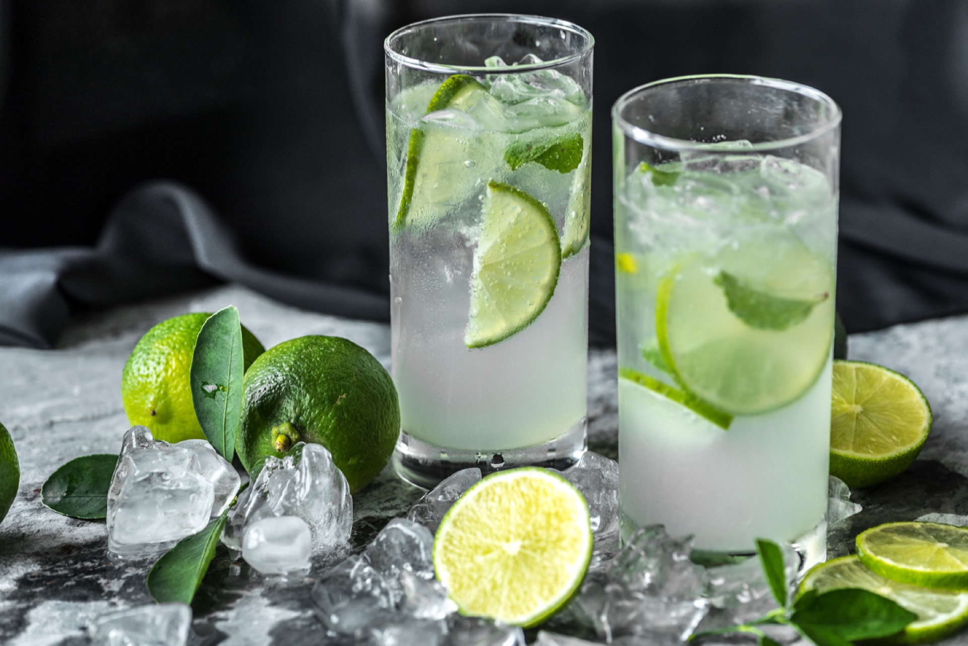 Two tall highball glasses of a clear drink over crushed ice with lime and mint, beside fresh limes